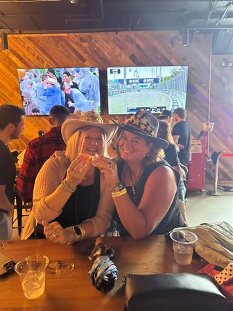 Two smiling women in cowboy hats sharing a snack at a lively sports bar with wooden accent wall and large TVs showing racing and crowd scenes