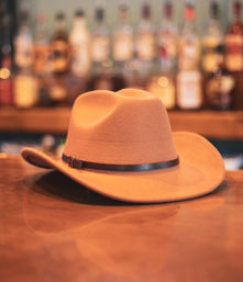 Brown felt cowboy hat with a black leather band resting on a wooden bar counter with blurred liquor bottles and warm bokeh in the background