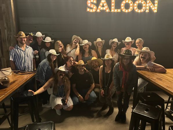 Group of about 18 people wearing cowboy hats and fake mustaches posing inside a dimly lit western-themed saloon with marquee "SALOON" sign, wooden tables and bar stools.