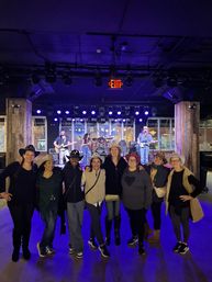 Eight friends in cowboy hats pose in front of a lit stage where a live country band plays under purple lights at an indoor music venue.