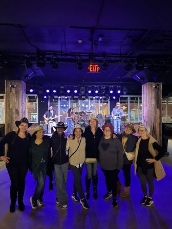 Eight friends in cowboy hats pose in front of a lit stage where a live country band plays under purple lights at an indoor music venue.