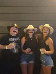 Three people wearing cowboy hats and fake mustaches pose with drinks in front of a dark wooden wall at a casual Western-themed party.