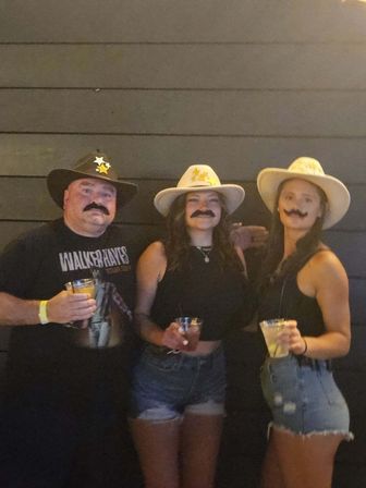 Three people wearing cowboy hats and fake mustaches pose with drinks in front of a dark wooden wall at a casual Western-themed party.