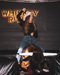 Smiling woman in a cowboy hat and jeans riding a mechanical bull indoors, one arm raised against a dark wood backdrop with warm ambient lighting.