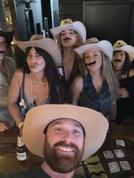 Western-themed bar group selfie — bearded man in the foreground and friends wearing cowboy hats and fake mustaches, posing and smiling over drinks on a lively night out