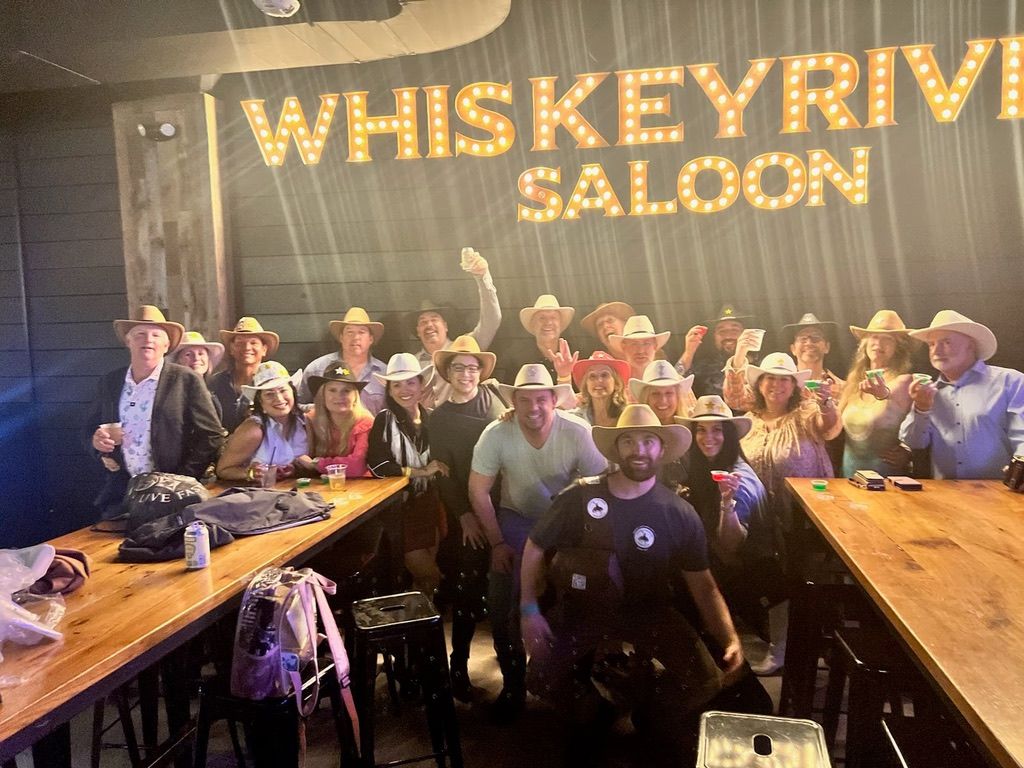 Large group wearing cowboy hats raising drinks in a lively western‑themed saloon under marquee lights with wooden tables and stools, upbeat nightlife scene.