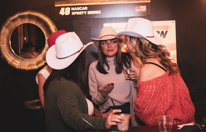 Four women wearing cowboy hats chatting and holding drinks at a dimly lit bar, cozy night out with friends.