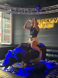 Person in a cowboy hat and fringe vest riding a mechanical bull inside a neon-lit saloon-style bar — playful country-night entertainment and indoor nightlife scene.