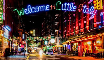 Vibrant Little Italy street at night with glowing 'Welcome to Little Italy' neon sign, neon-lit restaurants and cafes, outdoor seating and festive street lights.