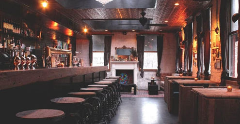 Cozy vintage-style bar interior with a long wooden counter and row of round stools, brass beer taps, exposed brick walls, candles on wooden booths, a decorative fireplace and tall curtained windows under warm ambient lighting.