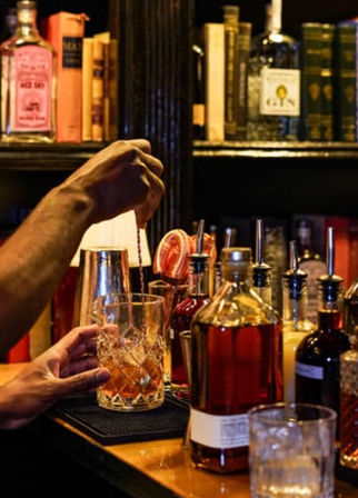 Hands stirring a classic whiskey cocktail in a cut-glass mixing glass on a cozy speakeasy-style cocktail bar counter, surrounded by whiskey bottles and bitters