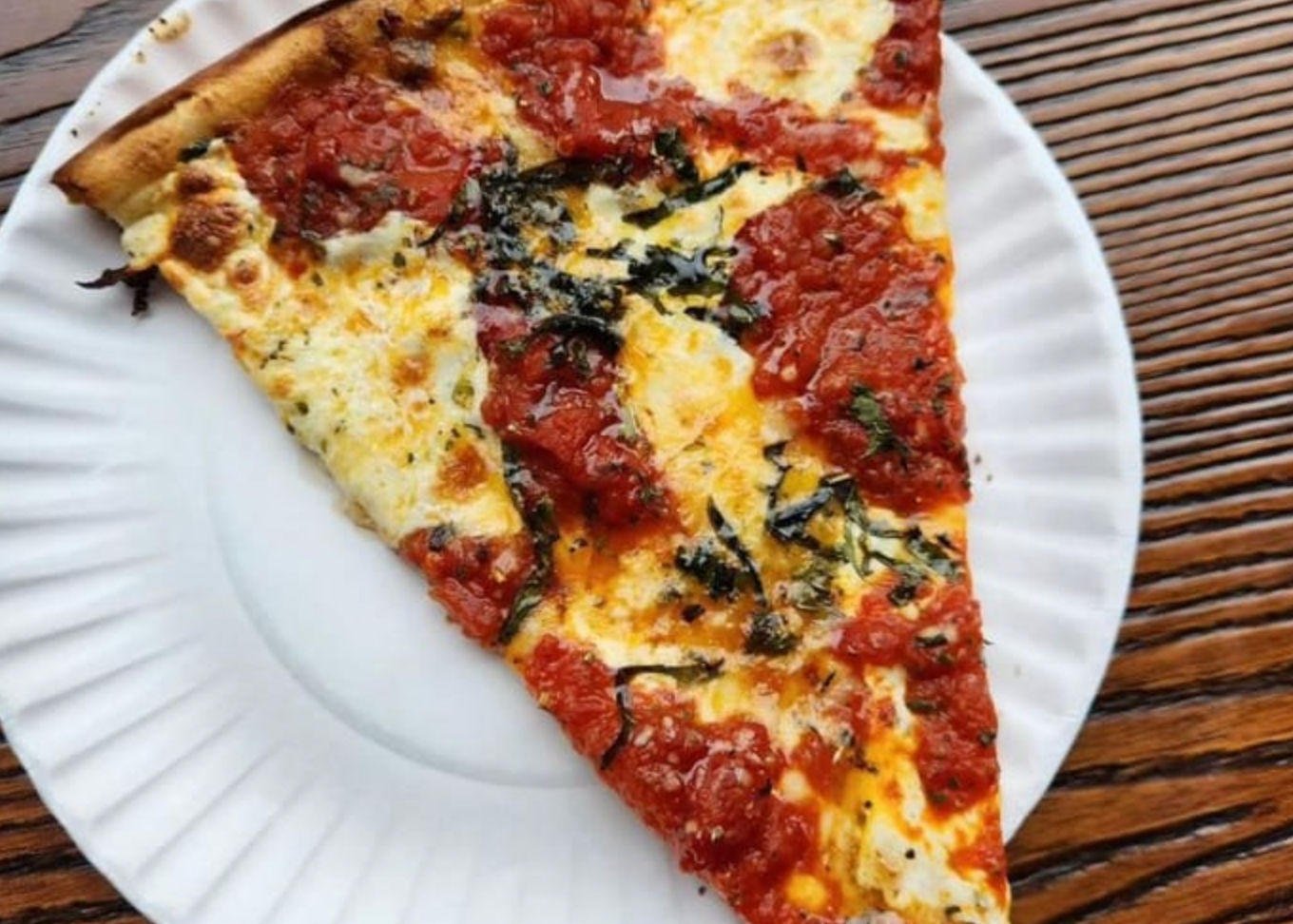Large margherita pizza slice with chunky tomato sauce, gooey mozzarella and fresh basil on a white paper plate over a wooden table