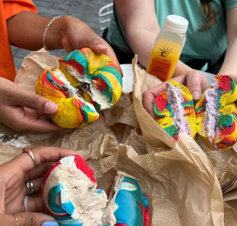 Three vibrant rainbow swirl bagels split and filled with cream cheese, held by multiple hands over crumpled brown paper on an outdoor table with a small juice bottle