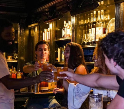 Group of friends toasting cocktails and laughing at a cozy, dimly lit cocktail bar with backlit liquor shelves — lively urban nightlife scene.