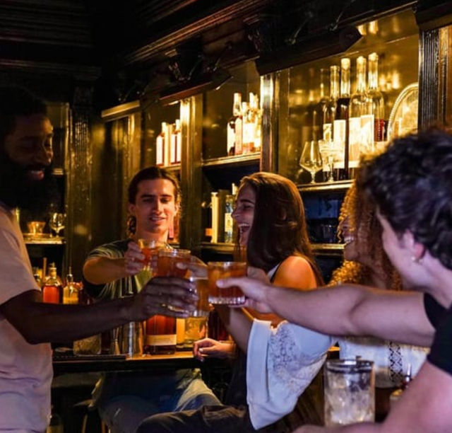 Group of friends toasting cocktails and laughing at a cozy, dimly lit cocktail bar with backlit liquor shelves — lively urban nightlife scene.