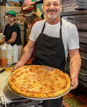 Smiling cook in a black apron holding a large golden cheese pizza on a metal tray in a busy neighborhood pizzeria kitchen.