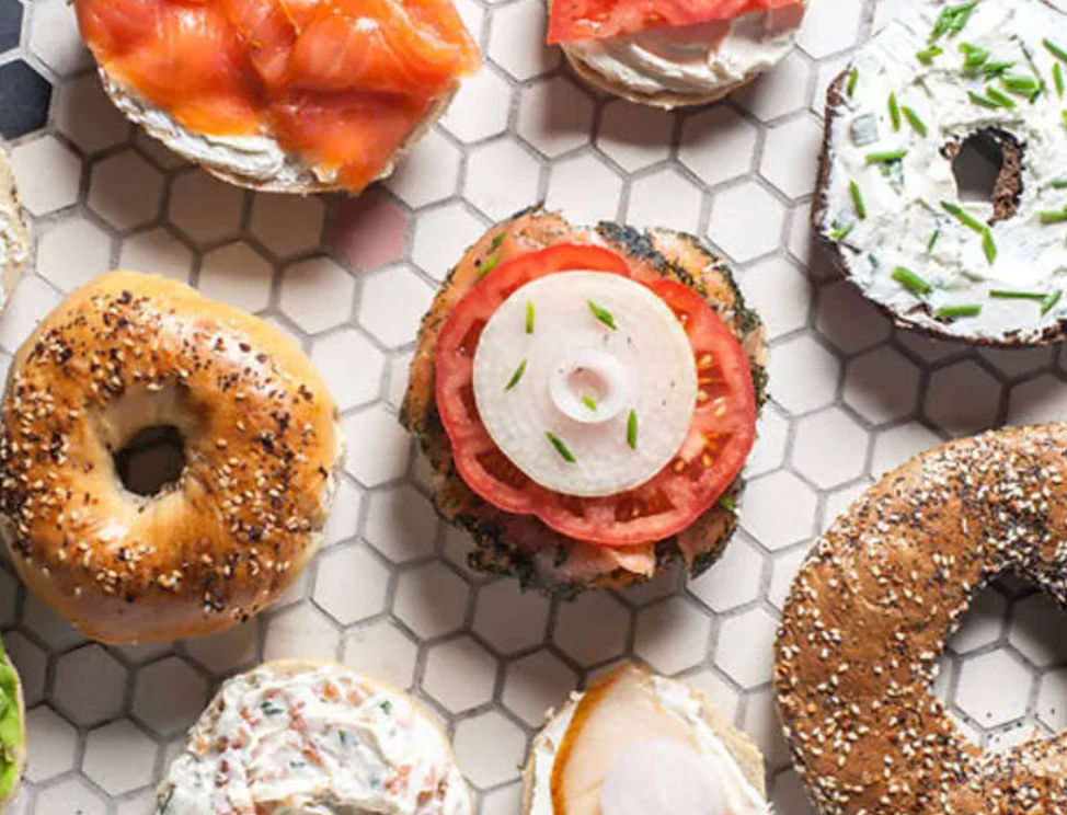 Assorted artisan bagels and halves topped with cream cheese, smoked salmon, sliced tomato and onion arranged on white hexagon tile — colorful brunch spread.