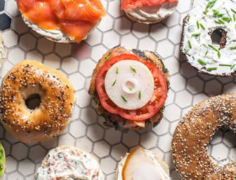 Assorted artisan bagels and halves topped with cream cheese, smoked salmon, sliced tomato and onion arranged on white hexagon tile — colorful brunch spread.