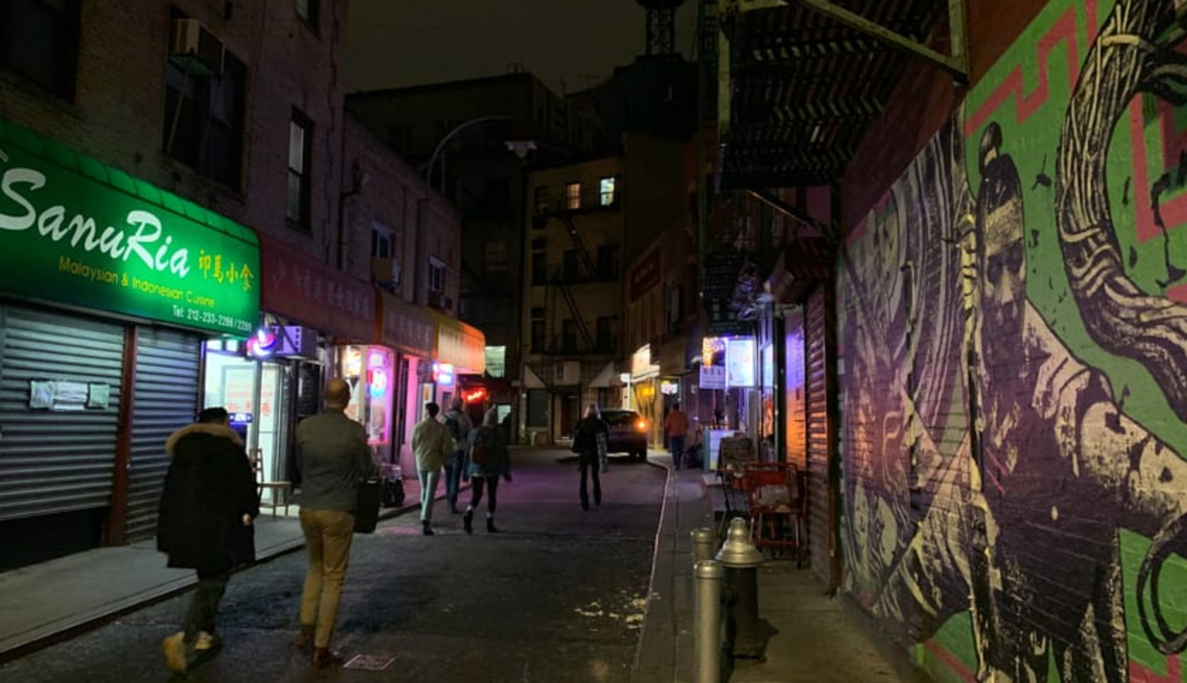 Vibrant night city alley with neon-lit restaurant storefronts, pedestrians walking under fire escapes, and a bold colorful street-art mural on the brick wall.