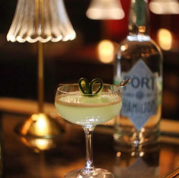 Pale green cocktail in a coupe glass topped with a curled cucumber garnish, placed on a polished bar beside a warm table lamp and a blurred spirit bottle — moody upscale bar scene.