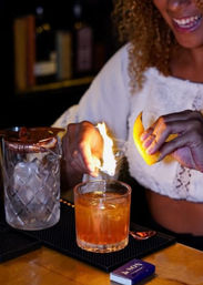 Hands torching a lemon peel to flame-spritz an amber cocktail in a rocks glass on a bar mat, with an ice-filled mixing glass and matches nearby.