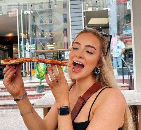 Smiling young woman at an outdoor city cafe taking a big bite of a long, cheesy pizza slice, wearing sunglasses on her head and a smartwatch.