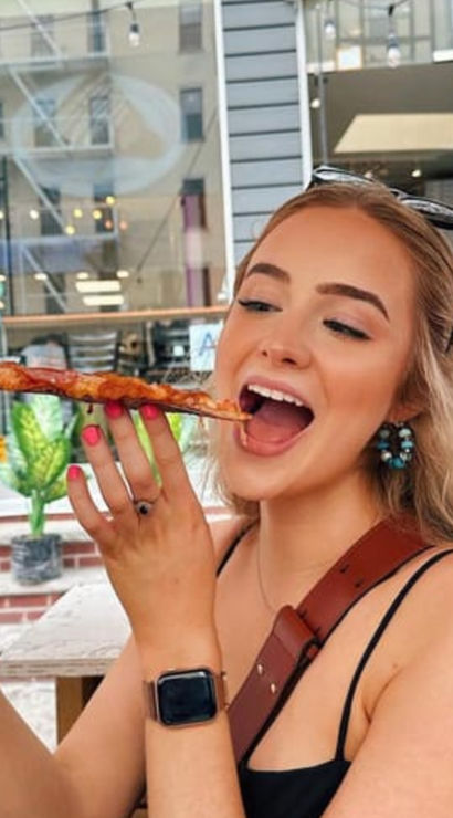 Smiling young woman at an outdoor city cafe taking a big bite of a long, cheesy pizza slice, wearing sunglasses on her head and a smartwatch.