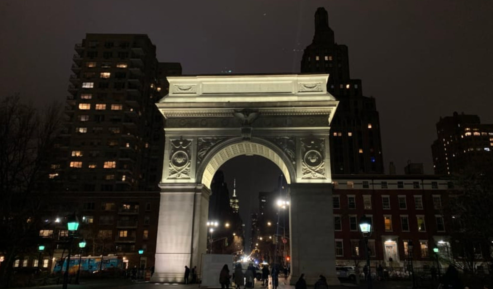 Illuminated Washington Square Arch at night in New York City, pedestrians walking beneath the glowing arch framed by apartment buildings and street lamps.