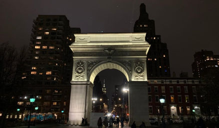 Illuminated Washington Square Arch at night in New York City, pedestrians walking beneath the glowing arch framed by apartment buildings and street lamps.