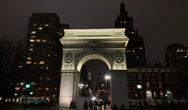 Illuminated Washington Square Arch at night in New York City, pedestrians walking beneath the glowing arch framed by apartment buildings and street lamps.