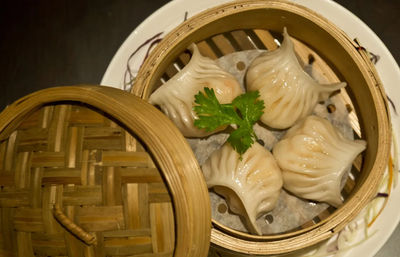 Close-up of four glossy pleated steamed dumplings (dim sum) in a round bamboo steamer basket on a white plate, garnished with a sprig of cilantro