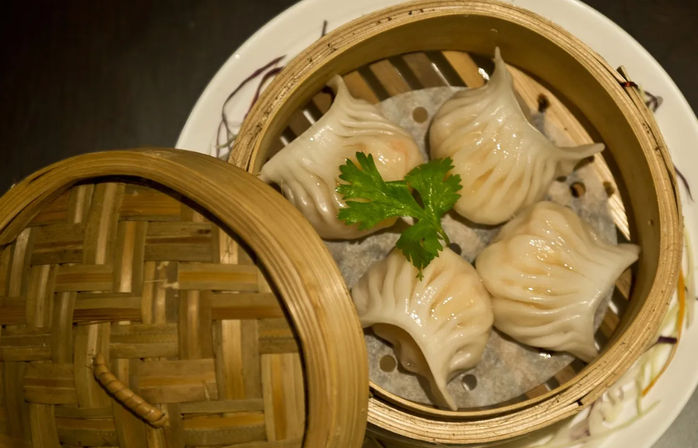 Close-up of four glossy pleated steamed dumplings (dim sum) in a round bamboo steamer basket on a white plate, garnished with a sprig of cilantro
