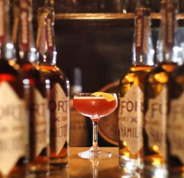 Amber-red cocktail in a coupe glass with an orange peel garnish, framed by whiskey bottles on a wooden bar in a cozy craft cocktail bar.