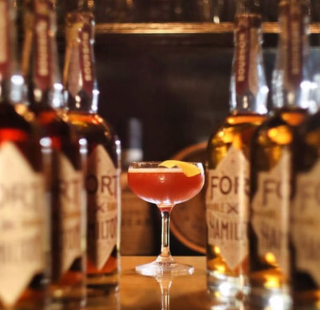 Amber-red cocktail in a coupe glass with an orange peel garnish, framed by whiskey bottles on a wooden bar in a cozy craft cocktail bar.