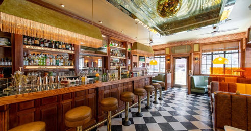 Retro downtown restaurant bar interior with long wood-paneled counter, leather swivel stools, black-and-white checkered floor, mirrored back bar stocked with bottles, gold disco ball and fringed pendant lamps over cozy booths.