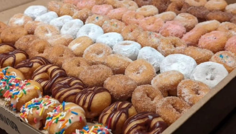 Assorted mini donuts neatly arranged in rows in a cardboard box — powdered sugar, cinnamon sugar, chocolate-drizzled and sprinkle-topped varieties, party-ready treat