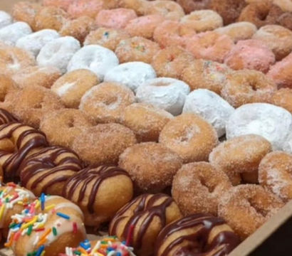 Assorted mini donuts neatly arranged in rows in a cardboard box — powdered sugar, cinnamon sugar, chocolate-drizzled and sprinkle-topped varieties, party-ready treat