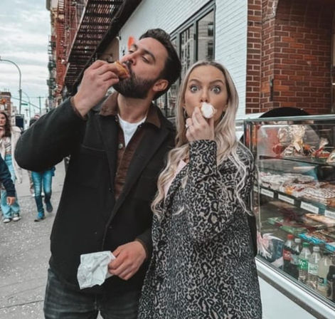Man and woman on a city sidewalk outside a bakery, both eating cream-filled pastries — woman in a leopard-print coat makes a playful surprised face next to a pastry display case and brick storefront.