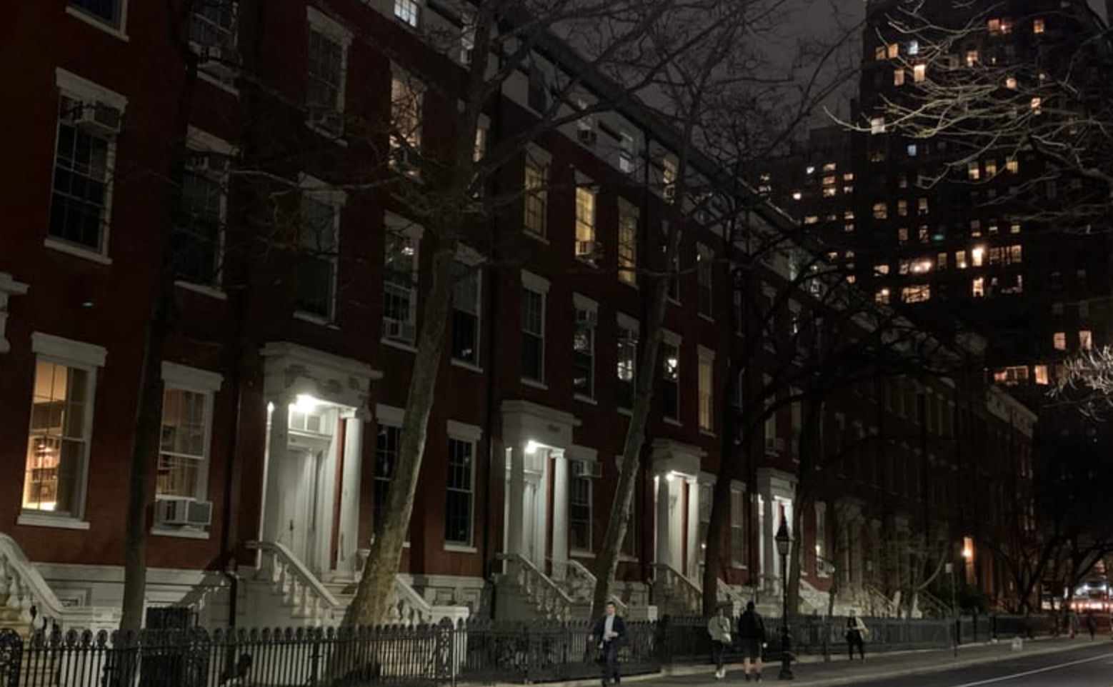 Nighttime city street of red brick brownstone rowhouses with white stoops, leafless trees and iron fence, illuminated windows and a distant high-rise, pedestrians on the sidewalk.