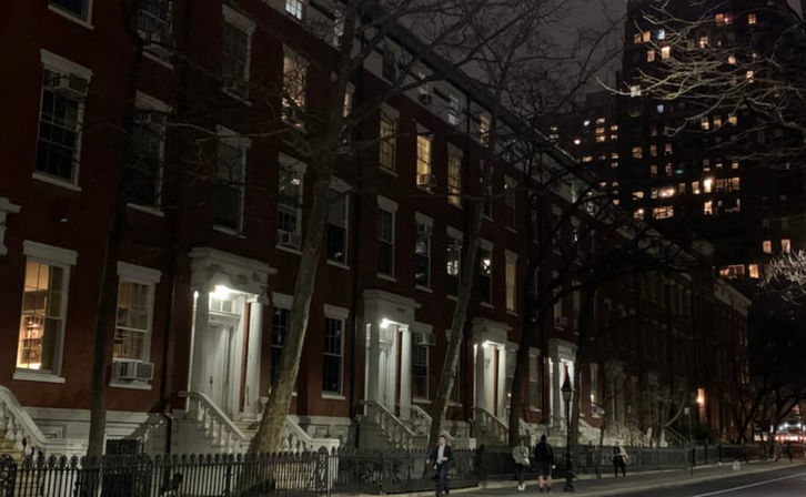 Nighttime city street of red brick brownstone rowhouses with white stoops, leafless trees and iron fence, illuminated windows and a distant high-rise, pedestrians on the sidewalk.
