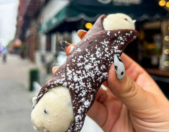 Chocolate-dipped, cream-filled cannoli dusted with powdered sugar held by a hand with black-and-white nail art, blurred urban street and sidewalk in the background