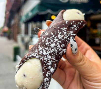Chocolate-dipped, cream-filled cannoli dusted with powdered sugar held by a hand with black-and-white nail art, blurred urban street and sidewalk in the background