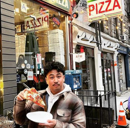 Person enjoying a cheesy slice of pizza on a paper plate outside a city pizzeria storefront with a bright PIZZA sign, stacked pizza boxes in the window, ATM and sidewalk railings.