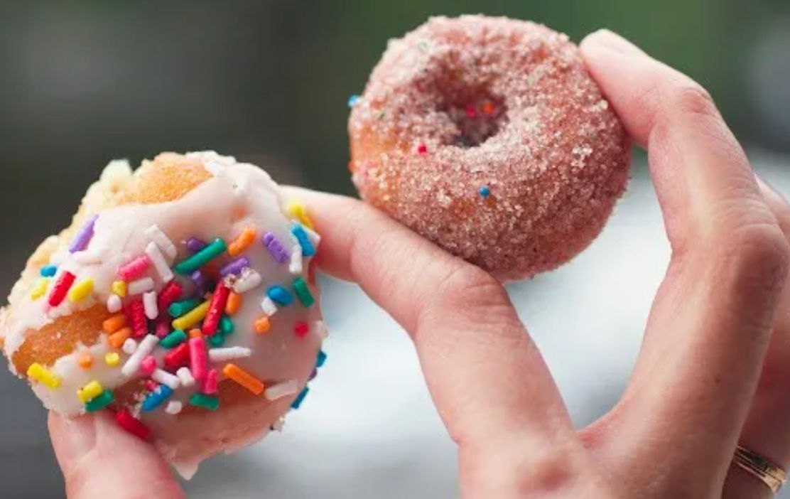 Close-up of two bite-sized mini donuts held in hand — one white-iced with rainbow sprinkles and one cinnamon-sugar coated
