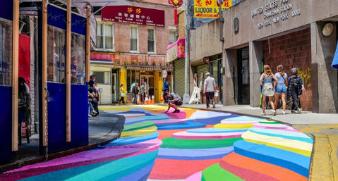 Vibrant rainbow-striped street mural winding through a Chinatown city block with pedestrians and storefronts — colorful urban crosswalk and street art.