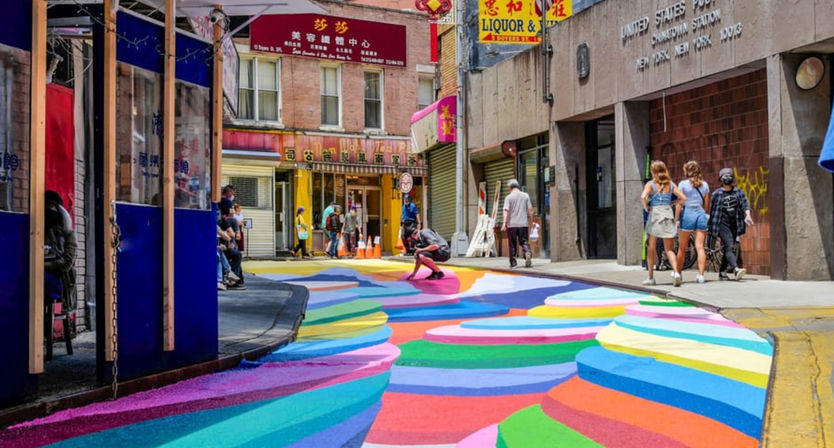 Vibrant rainbow-striped street mural winding through a Chinatown city block with pedestrians and storefronts — colorful urban crosswalk and street art.
