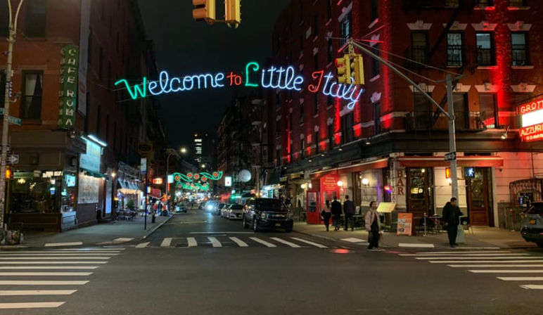Vibrant nighttime Little Italy, NYC street with glowing "Welcome to Little Italy" neon sign over a crosswalk, lit brick restaurants, parked cars and pedestrians.