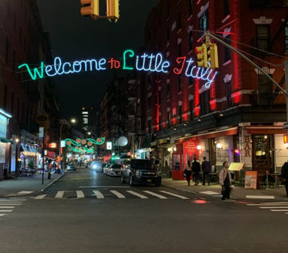 Vibrant nighttime Little Italy, NYC street with glowing "Welcome to Little Italy" neon sign over a crosswalk, lit brick restaurants, parked cars and pedestrians.