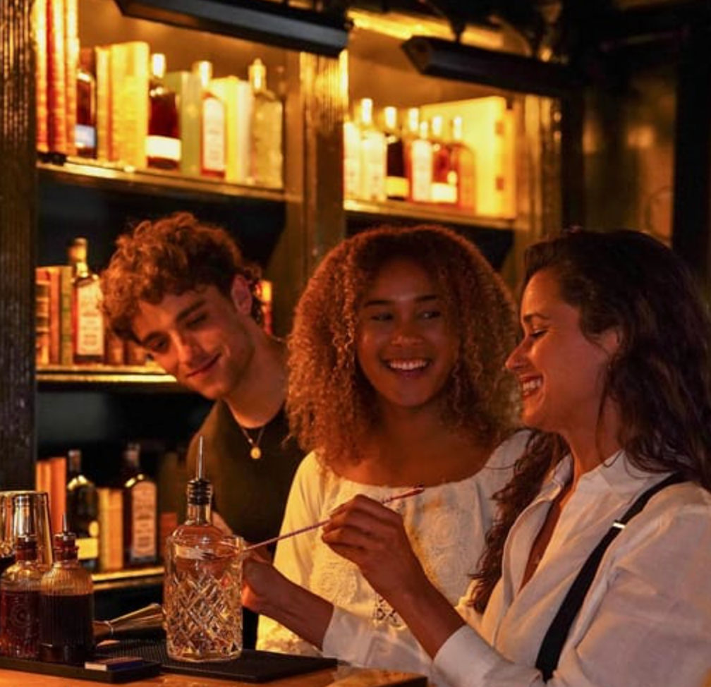 Three friends laughing at a cozy, dimly lit cocktail bar as one stirs a drink in a crystal mixing glass with shelves of liquor bottles behind them.