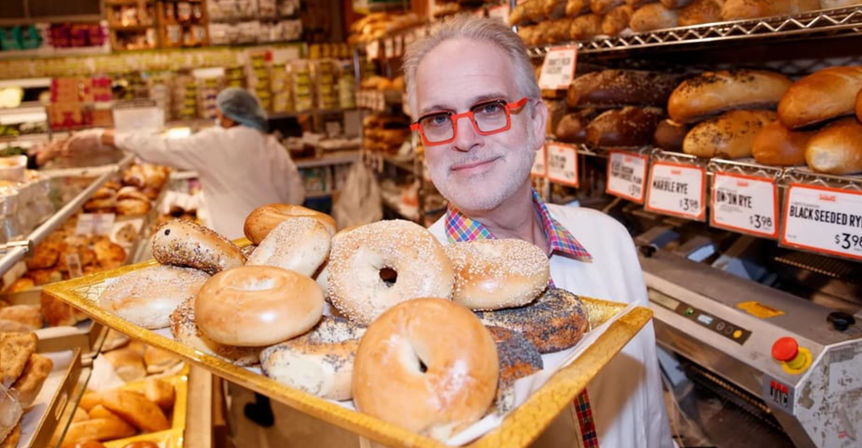 Smiling bakery employee holding a gold tray of assorted freshly baked bagels in a busy deli-style bakery with shelves of bread and price tags in the background.